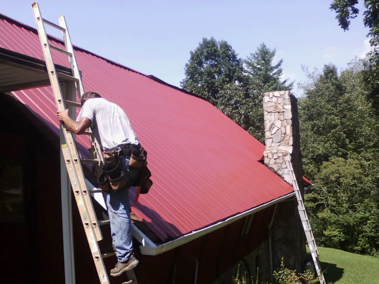 Roofer on a ladder beside a red metal roof and stone chimney — Soffit & Fascia Repair by Roofing Brothers in Martinsdale